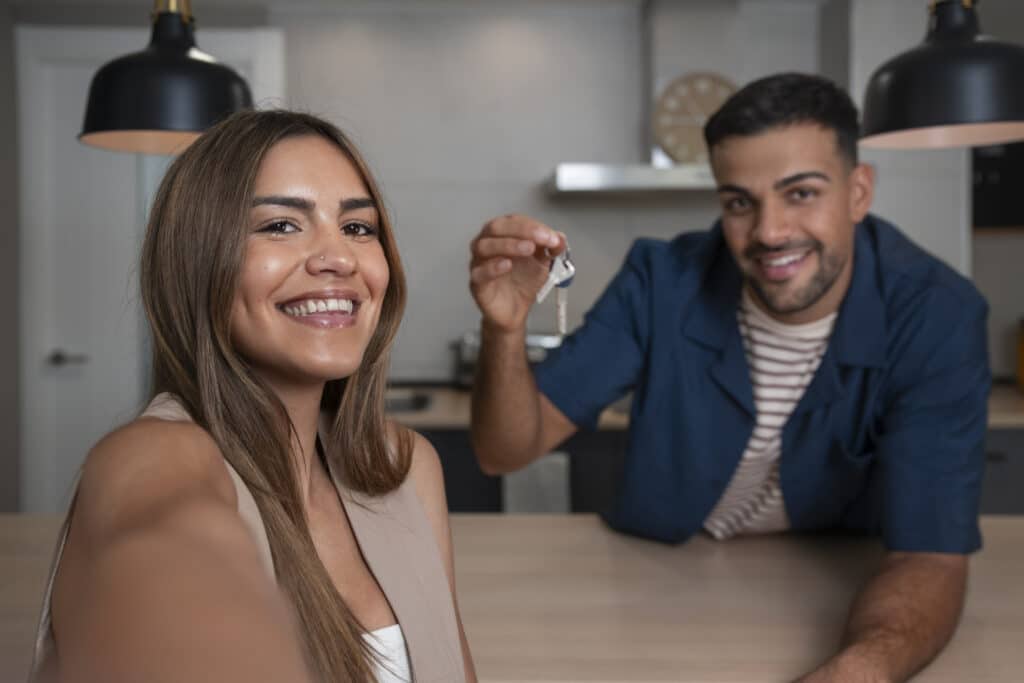 Man and woman smiling while taking a selfie inside their new home; man holding a house key.