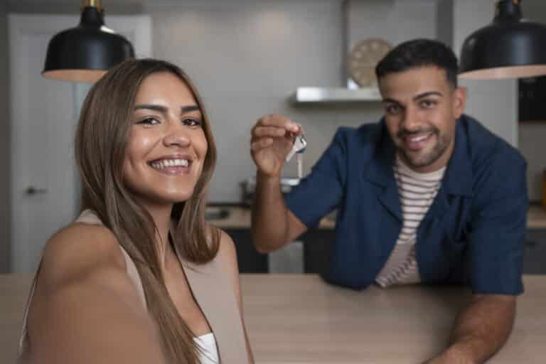 Man and woman smiling while taking a selfie inside their new home; man holding a house key.