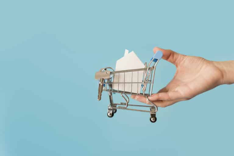 Mini shopping cart holding a small house, symbolizing buying a home, against a blue background